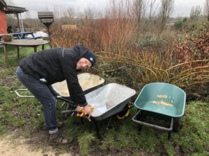 Photo of Simon, volunteering at Flower Pod Southwell. He is holding a pair of gardening shears and lifting a wheelbarrow.