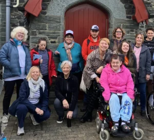 A group of people posing together outside a stone building with a red arched wooden door. The group includes individuals standing and seated, one person in a wheelchair wearing a bright pink jacket and blue trousers. Others are dressed in casual outdoor clothing such as jackets, scarves and trainers.