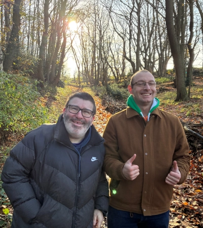 Man wearing black puffer coat smiling, stood next to man with glasses doing two thumbs up wearing brown jacket, both standing in a sunny forest